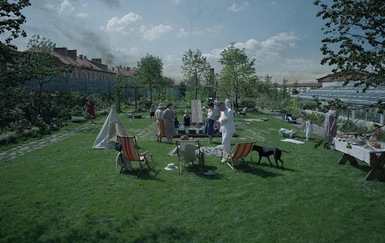 in an immaculate garden a family play in and around a small swimming pool. Beyond the garden wall, a barracks is visble with crematorium smoke rising beyond it.