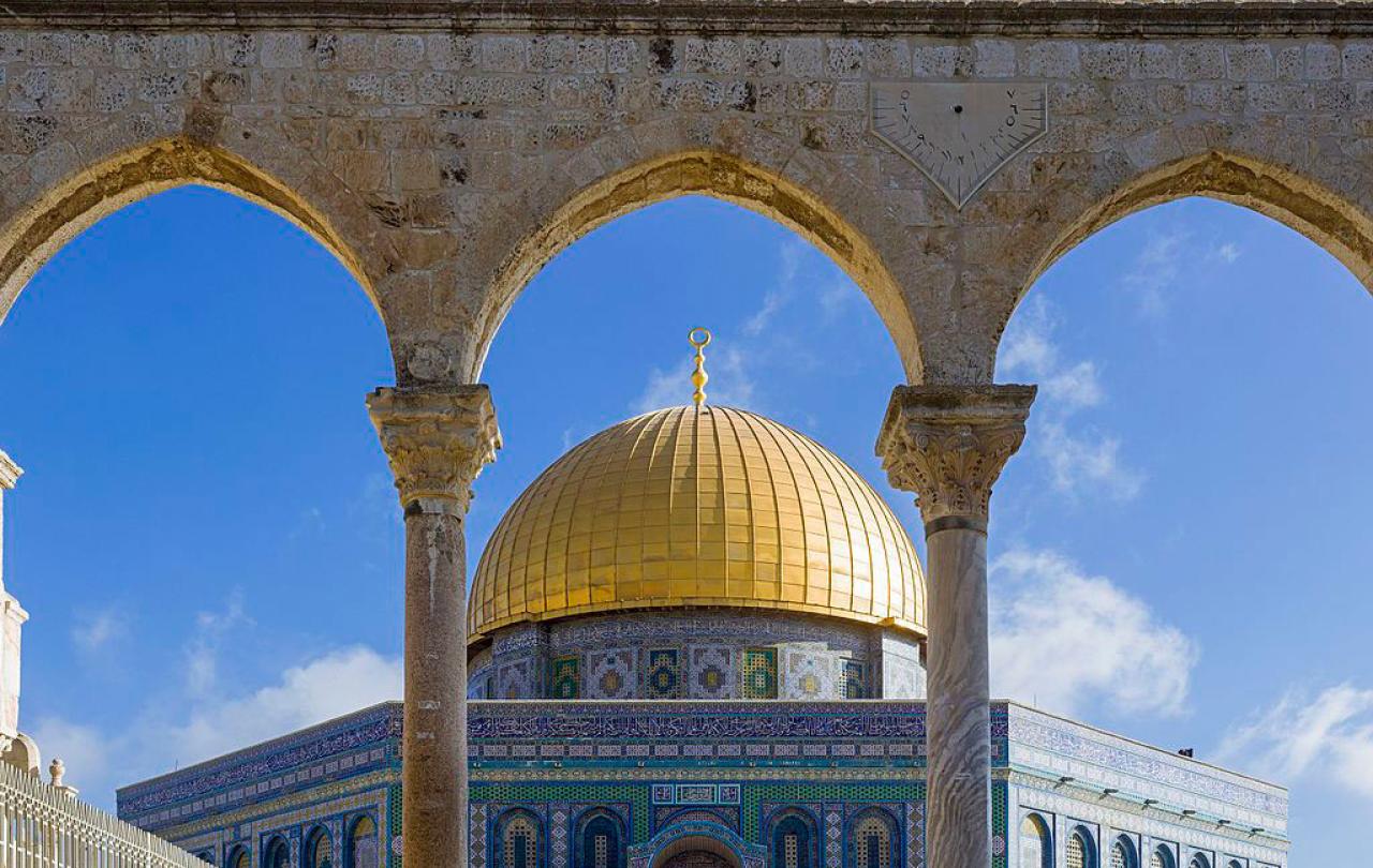 A gold-domed, blue-walled octagonal mosque seen through a row of arches.