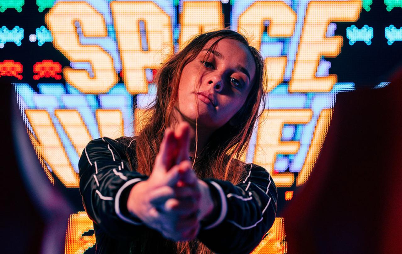 A woman stand in front of a large video screen displaying the Space Invaders title, hold her hands out in front of her.
