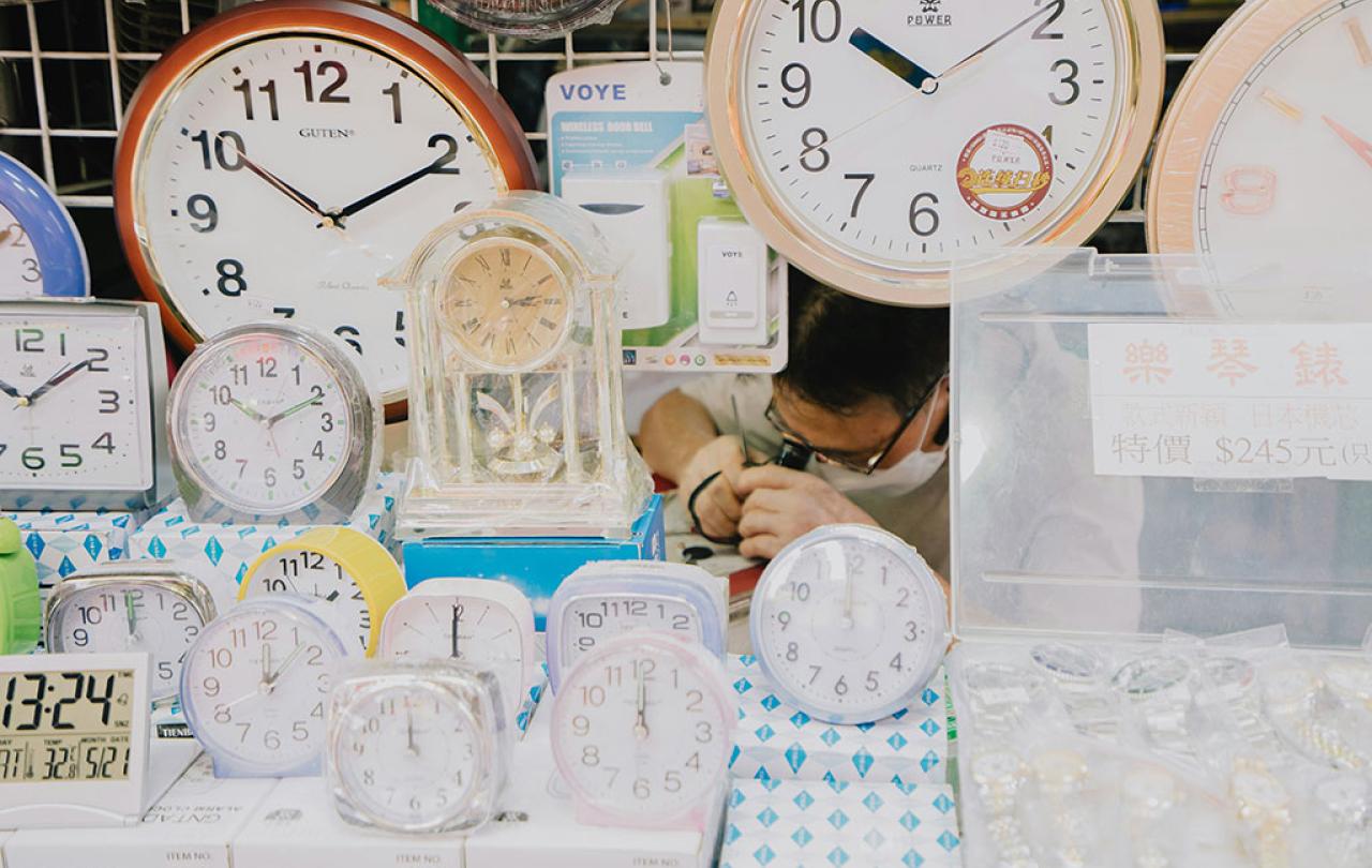 A clock repair peers at a clock he is repairing, amid a see of alarm and wall clocks on display