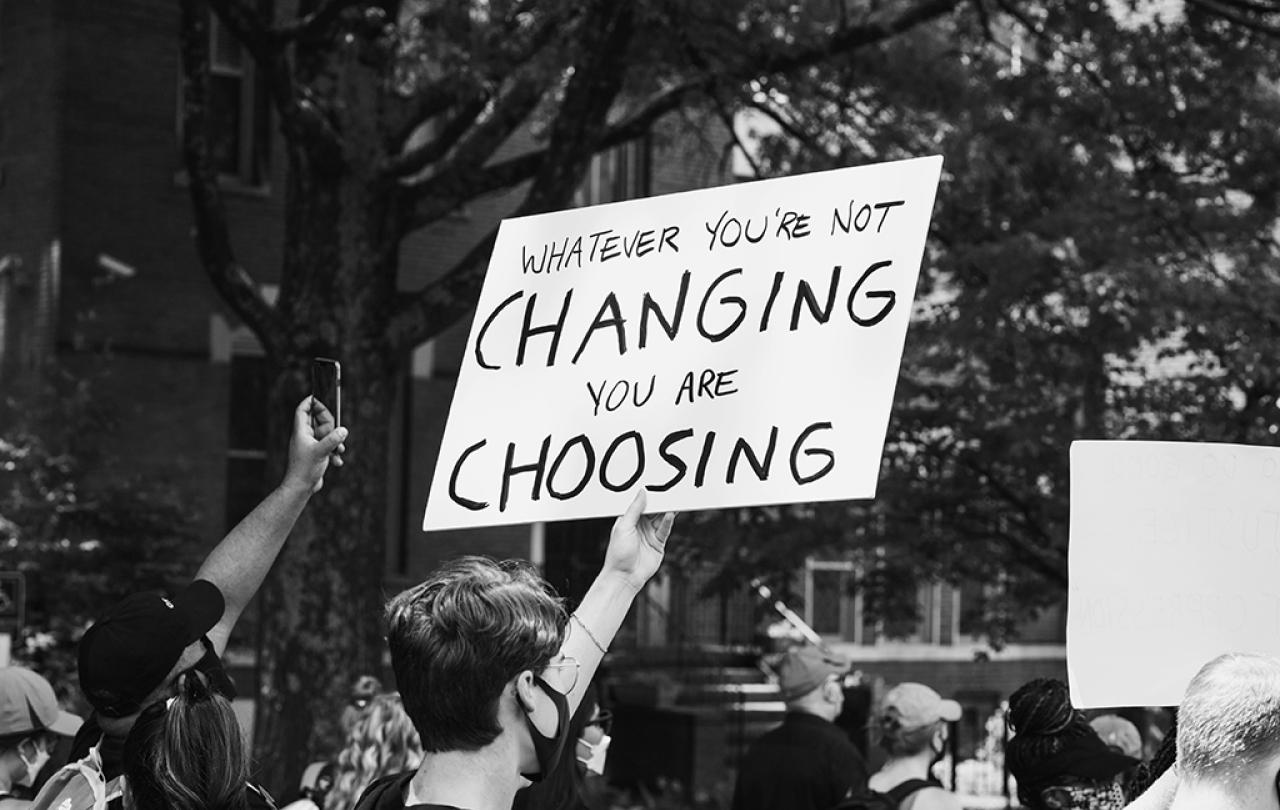 A protest placard is held above a march, reading 'If you don't change it, you choose it'.