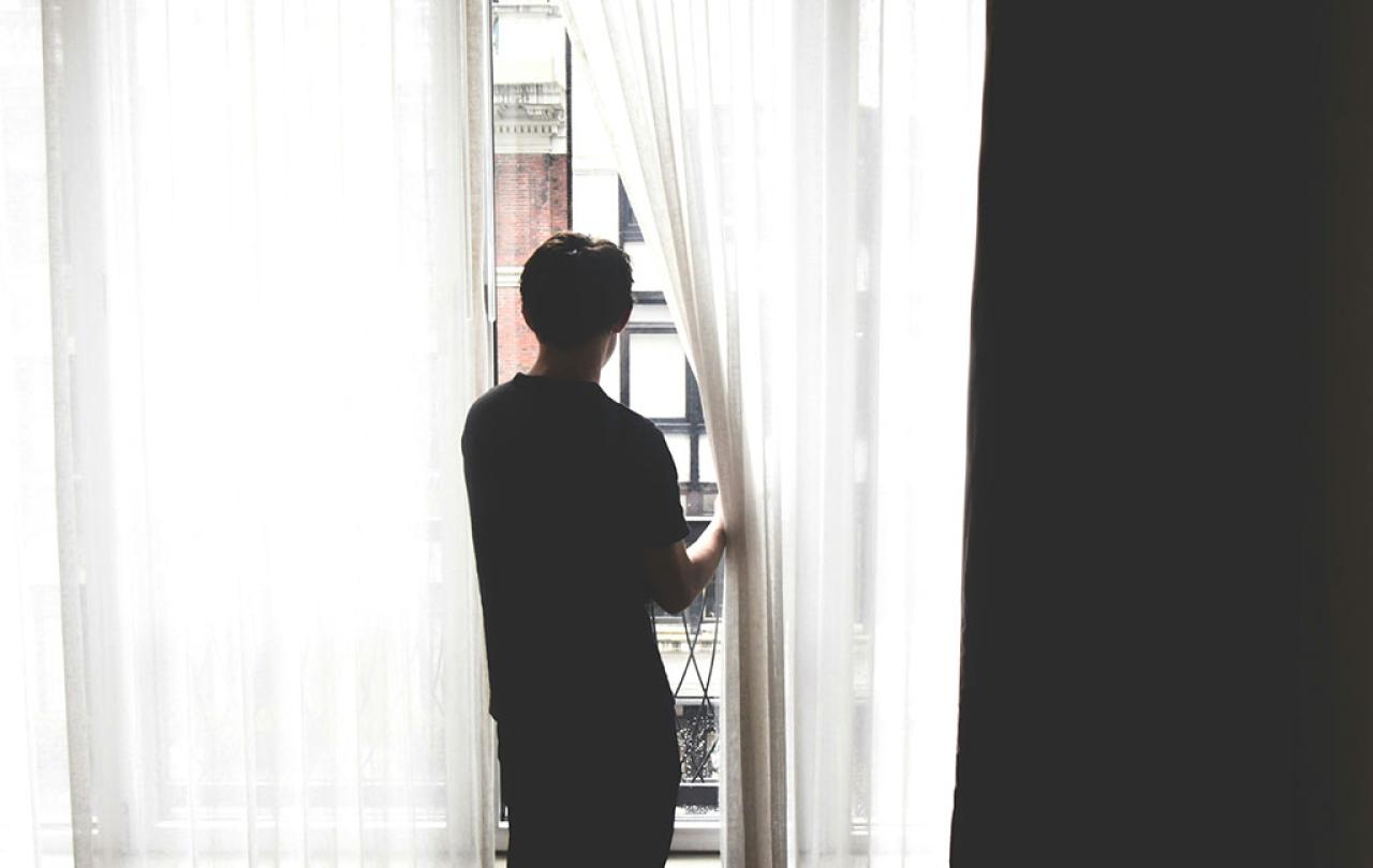 A persons stands, holding a net curtain aside to gaze out.