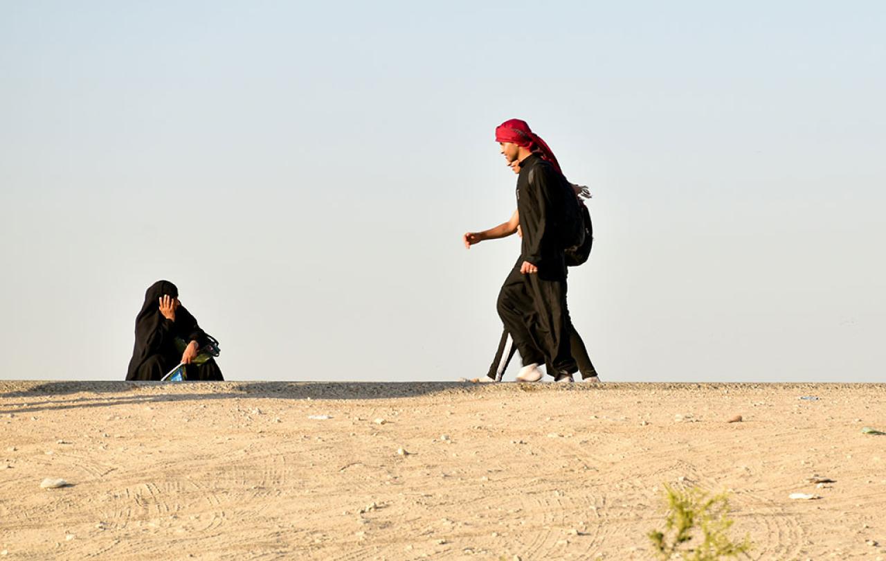 A black clad pilgrim squats and holds his head on the side of a desert road as other pilgrims walk along.