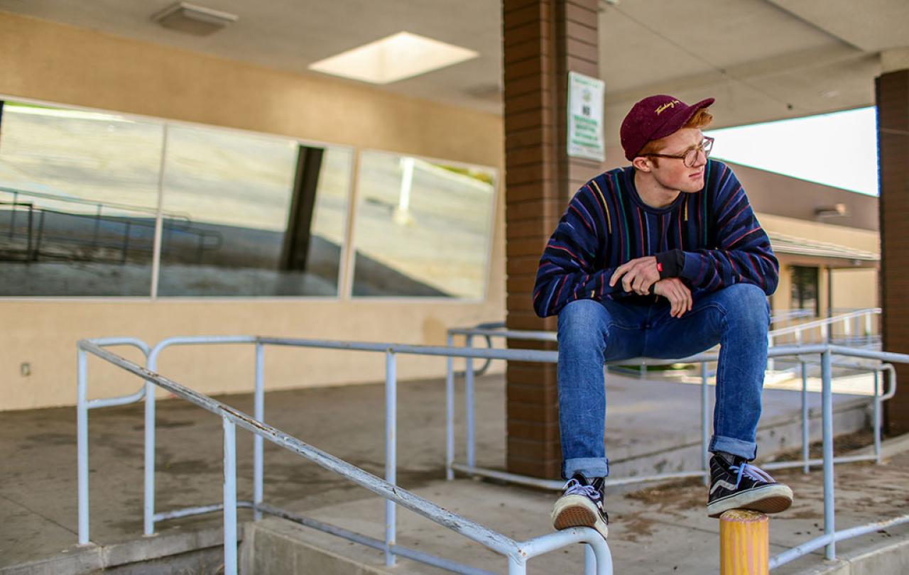 A casually dressed man perches on railing balancing, clasping his hands and looking around.