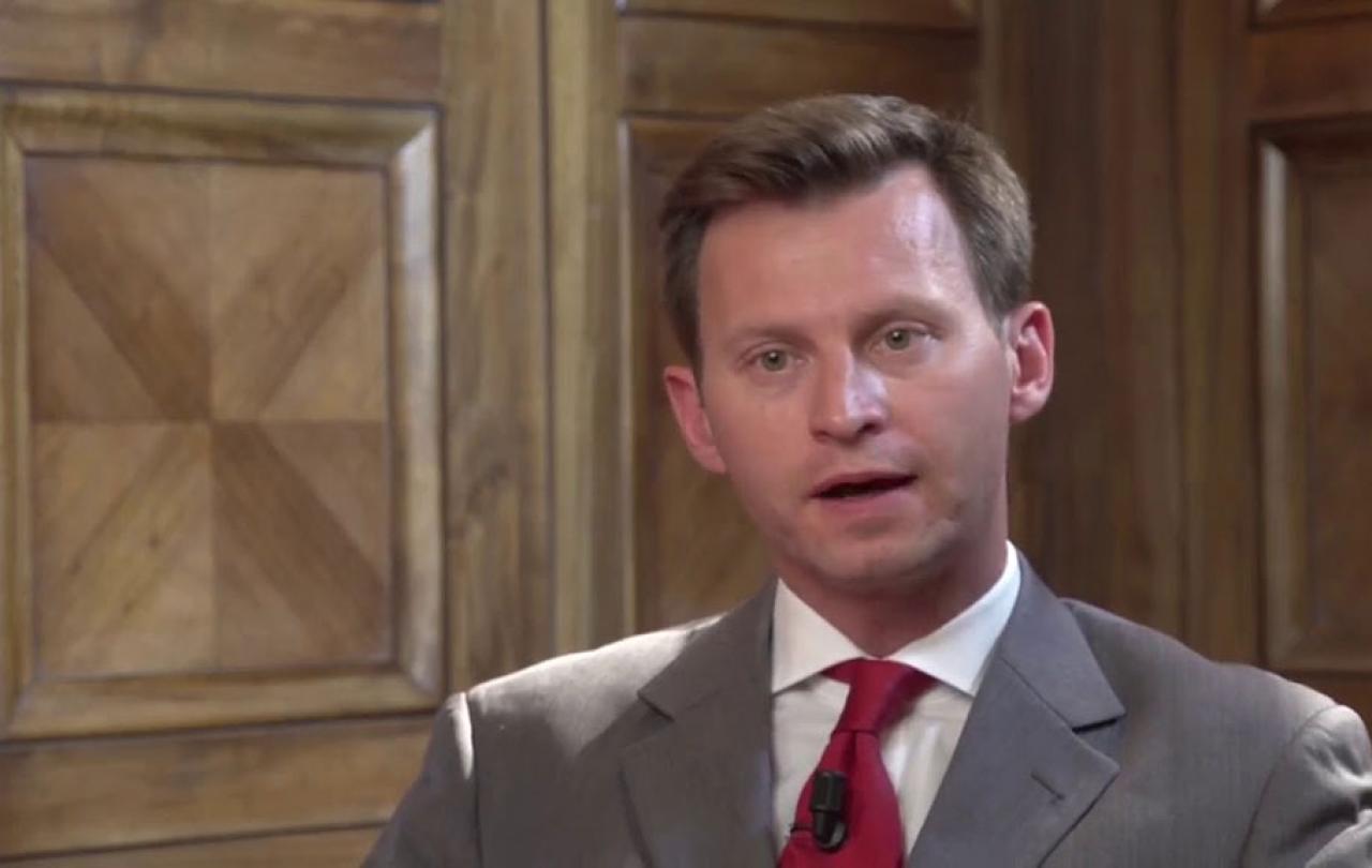 A man talks animatedly looking at the camera while sitting against a wood panelled wall.