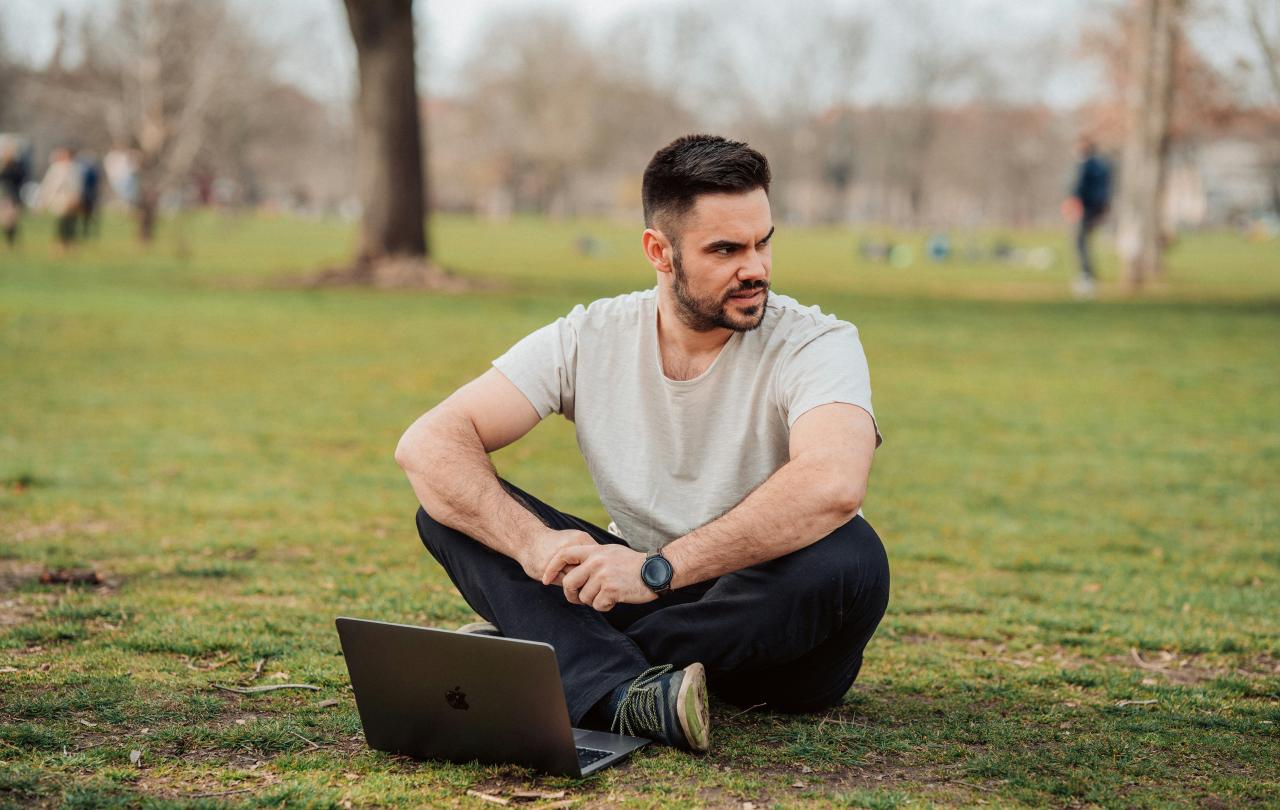 A man sits cross legged in a park with a laptop on the grass in front of him. He looks to one side.