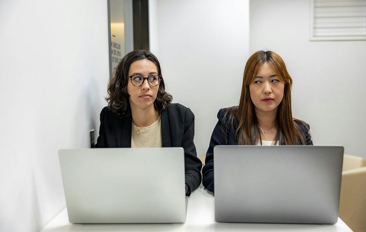 Two people sit beside each other working on laptops, One looks askance to the other who frowns.