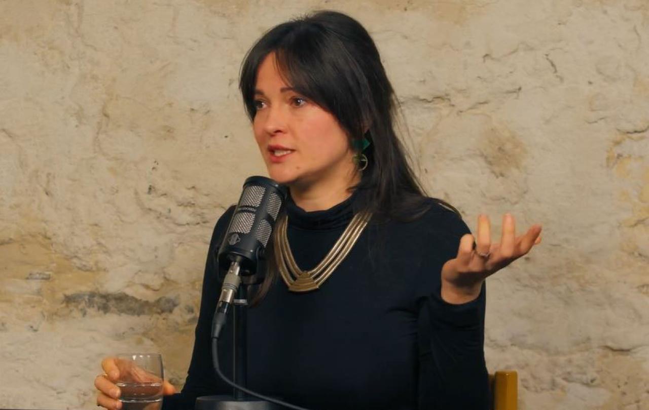 A woman sits a a table with a microphone in front of her, she is talking and gestures with one raise hand upward.