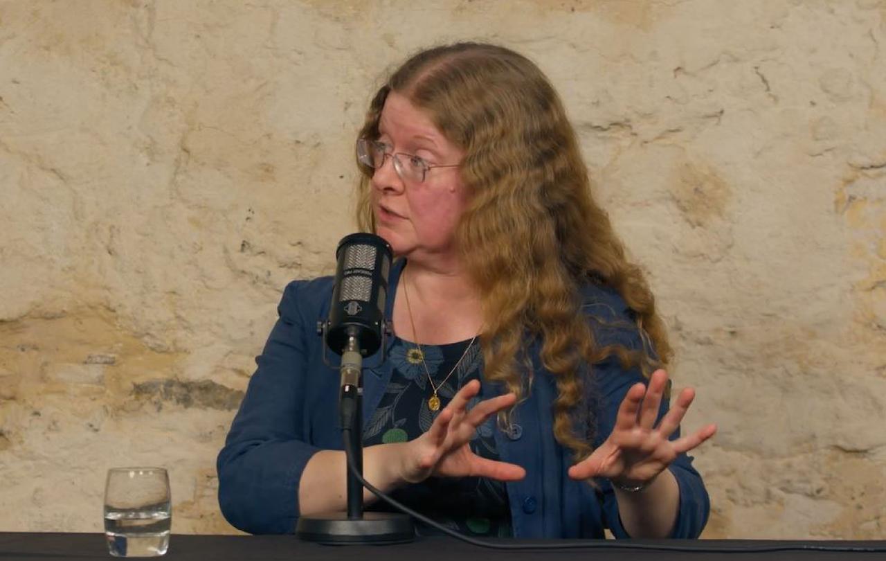 a woman seated in front a mic, looks to the left will gesturing both hands to the right.