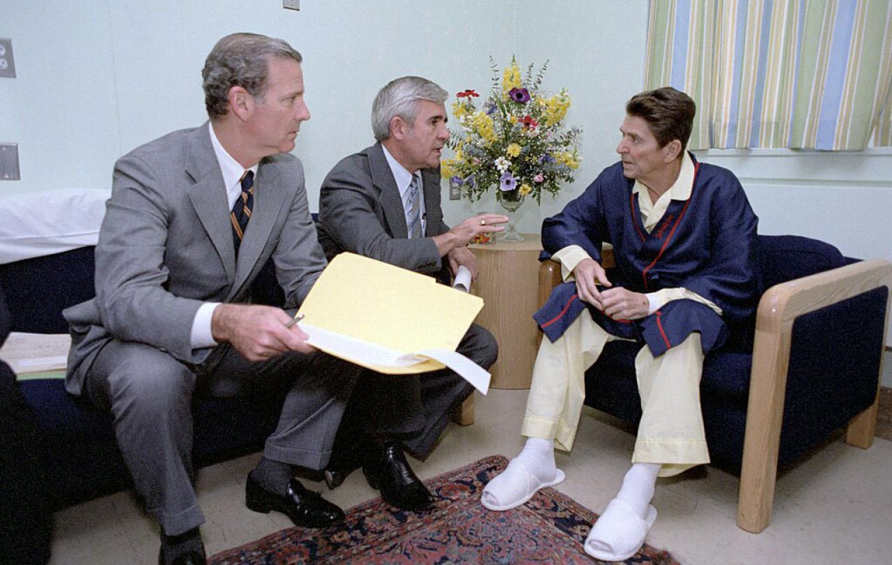 Two men in seats adress a pyjamap-clad Ronald Regan.