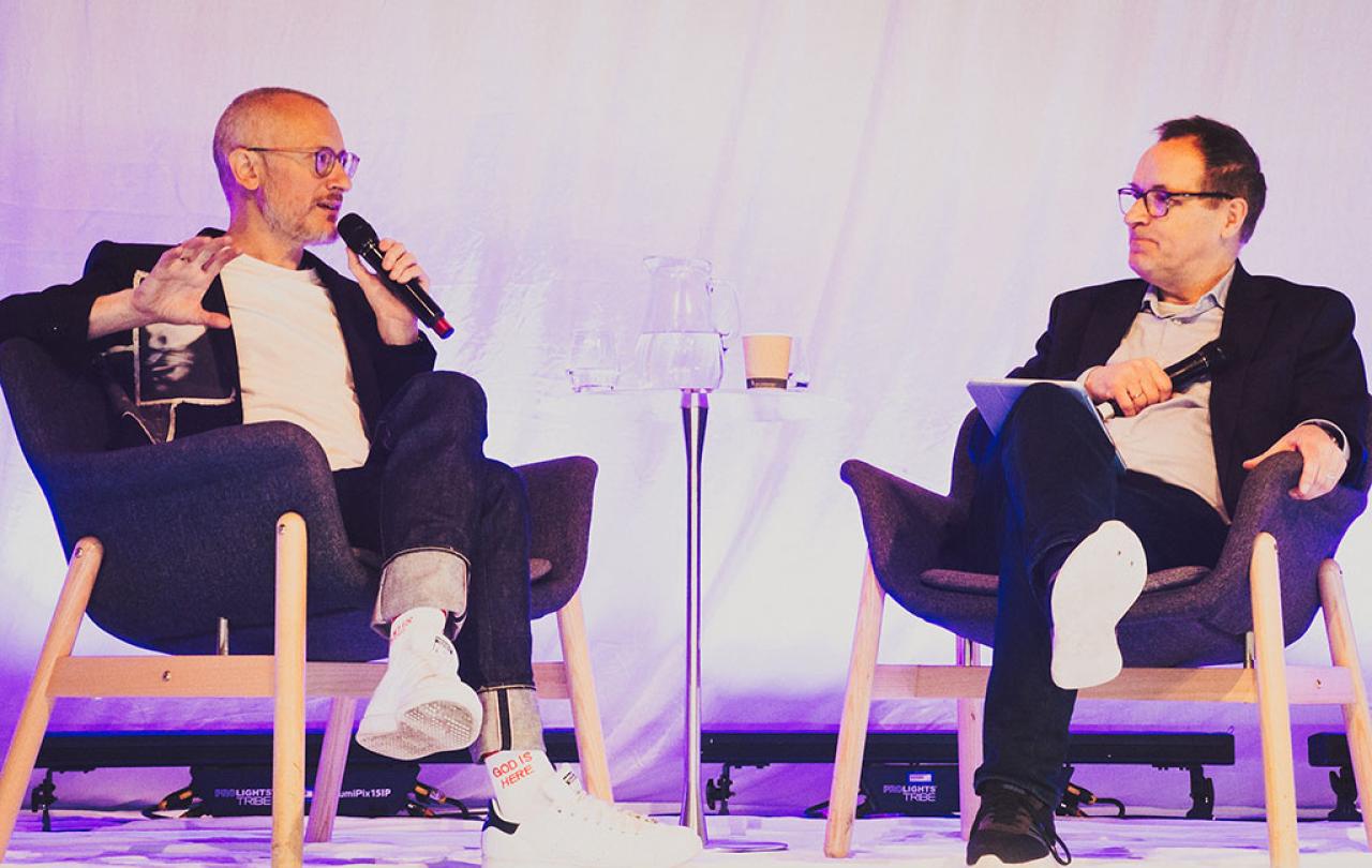 Two people sit on a stage in a relaxed manner, having a converation.