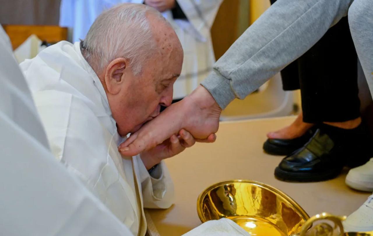 The Pope, wearing white, kneels, crades a bare foot, and kisses it.