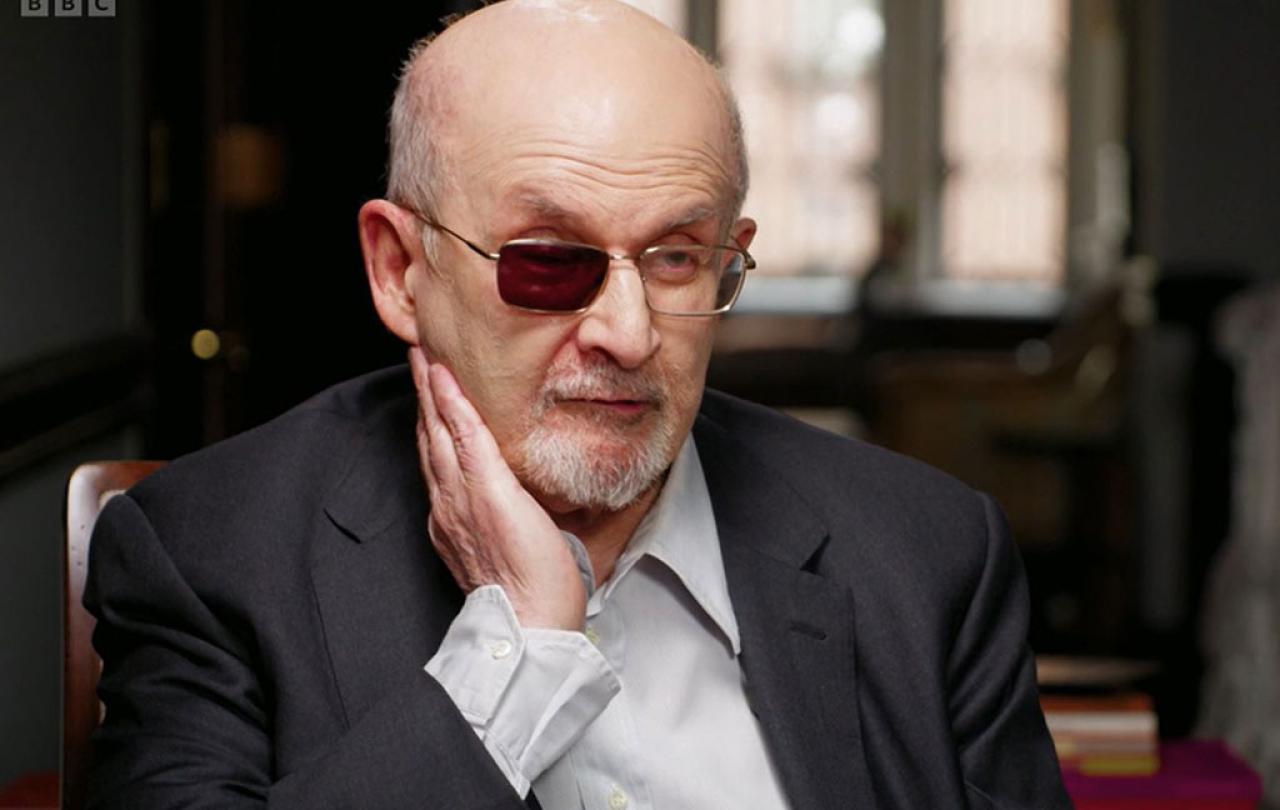 A man sits being interviewed and holds a hand to the side of his face, one lens of his glasses is tinted black.