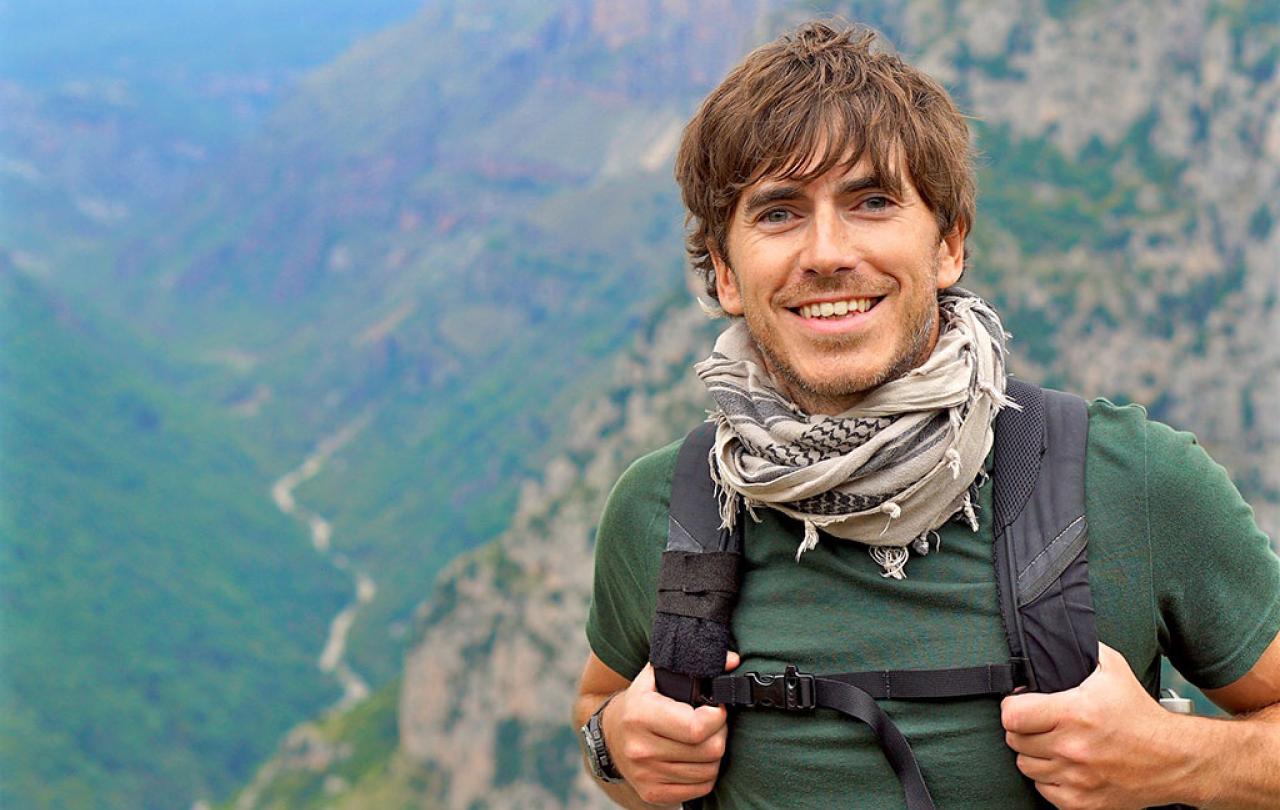 An enthusiastic hiker stands in front of a view down a valley, smiling and holding his backpack straps.