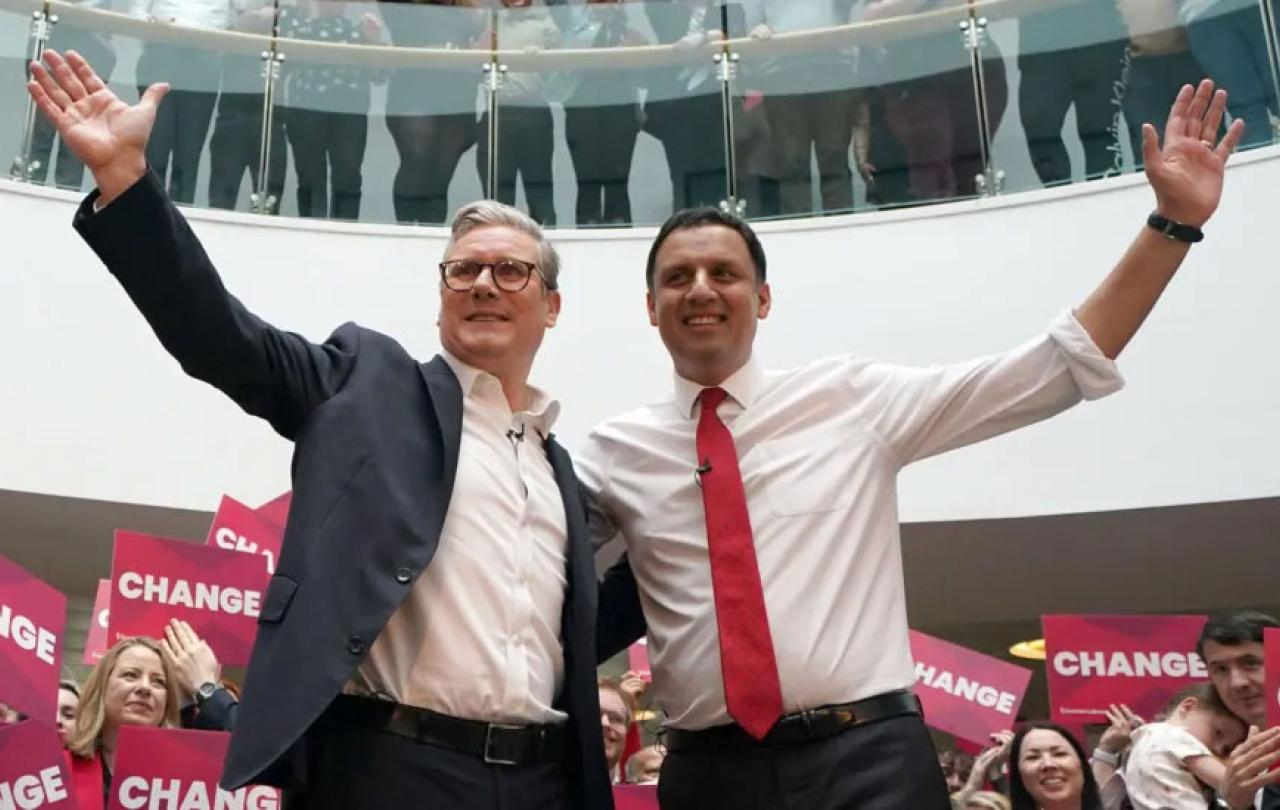 Two politicians, stand with one arm around each other and the other arms raised.