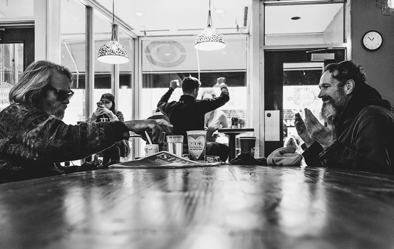 In a coffee shop, people sit around a table, one holds their hands up in surprise. 