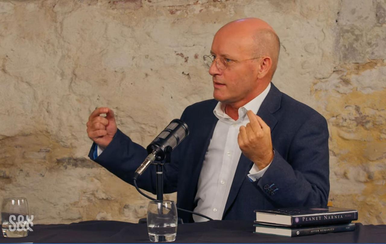 A man sits and speaks into a microphone and gestures with raised hands.