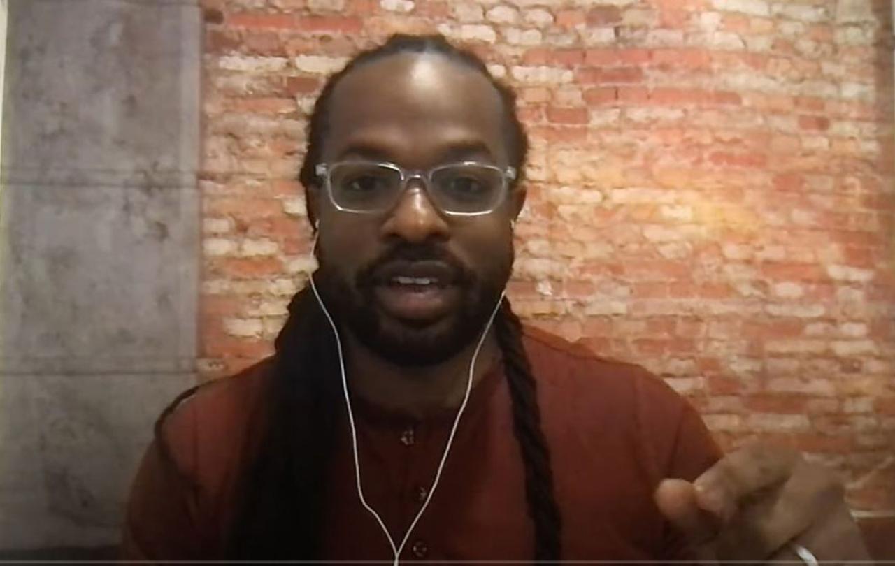 A man wearing white earphones talks to a camera and gestures with one hand.