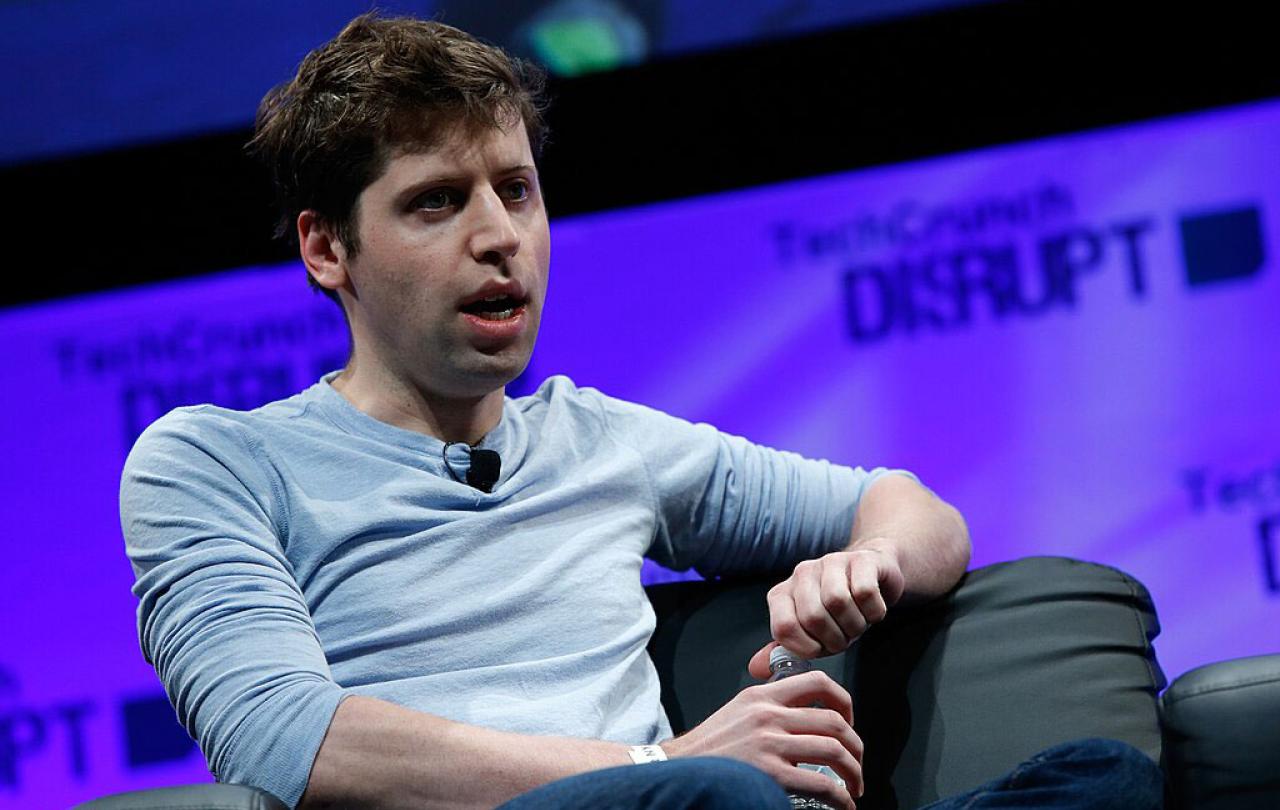On a conference stage, a seated speaker leans back and opines