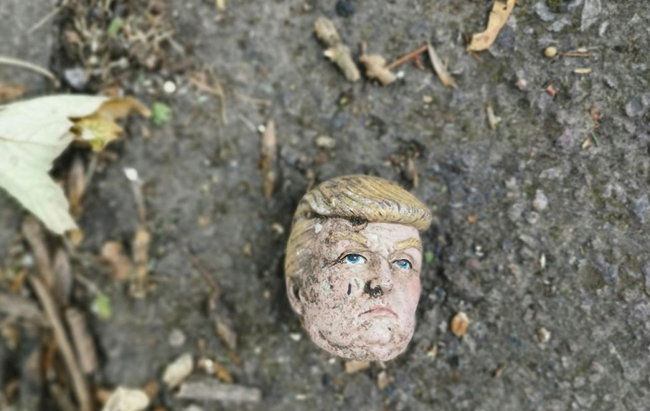 A severed doll head, resembling Donald Trump, lies on dirty ground.