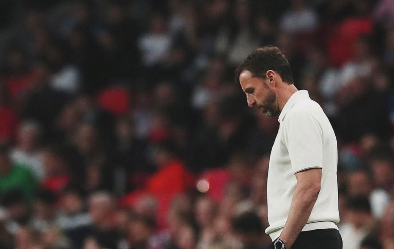 A dejected looking football manager ponders his feet while standing beside a pitch.