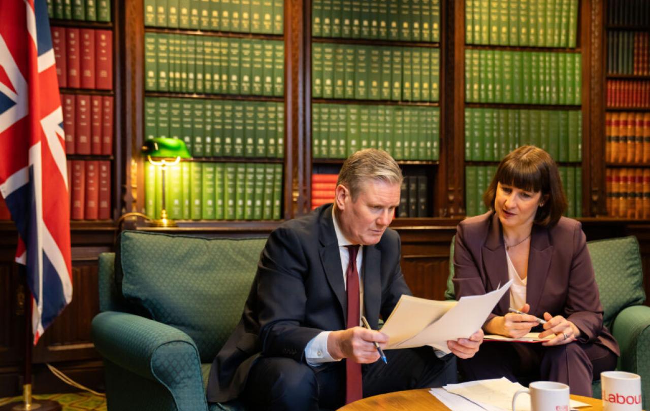 Kier Starmer sits on a sofa, leaning forward and holding papers he is reading. Rachel Reaves sits and looks on.