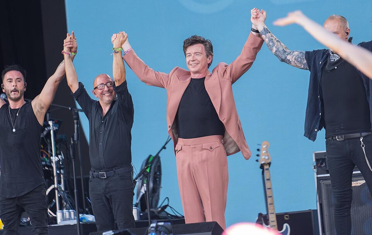 On a music festival stage, a popstar in a pink stages holds raised hands with his band.