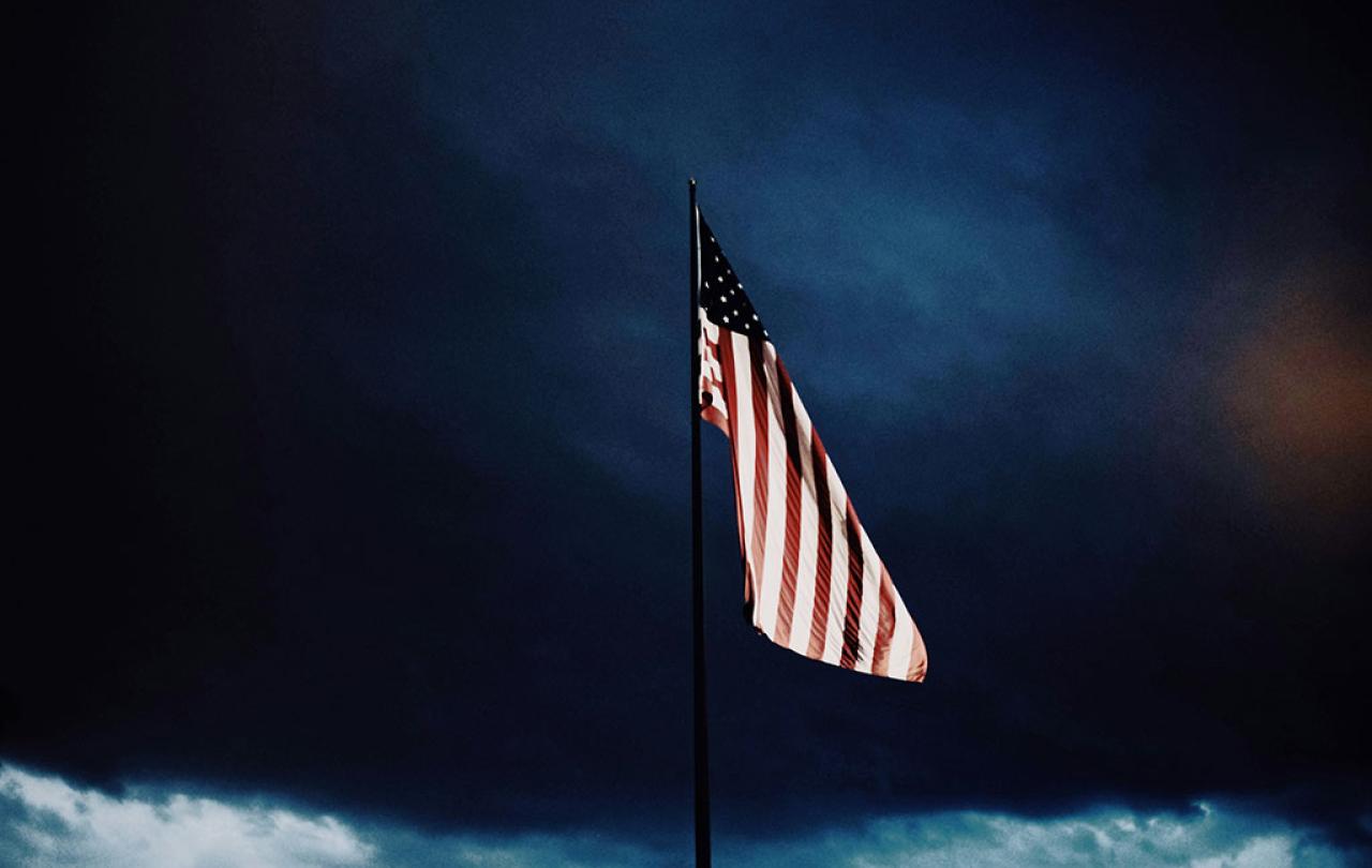 A US flag flutters under a dark brooding cloudy sky.