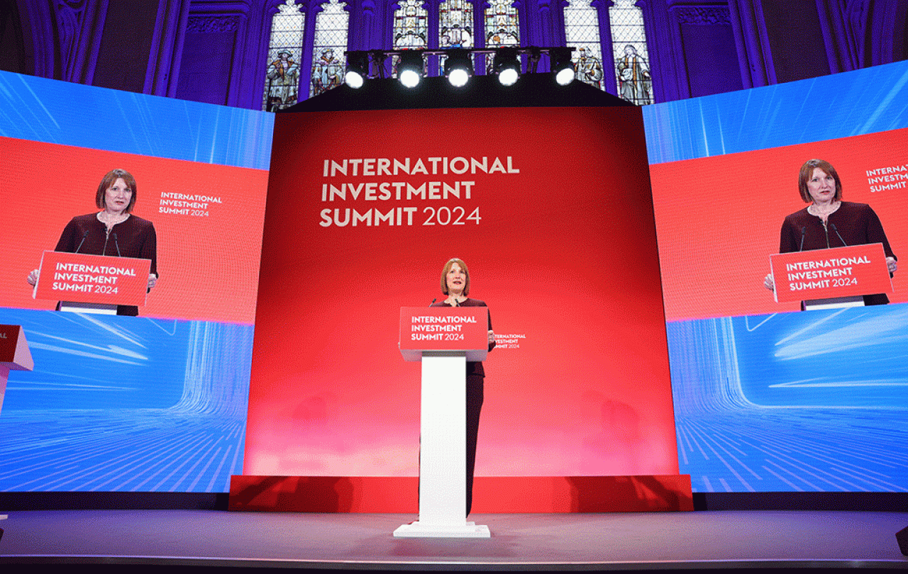 A woman stands behind a lectern against a blue and red backdrop.
