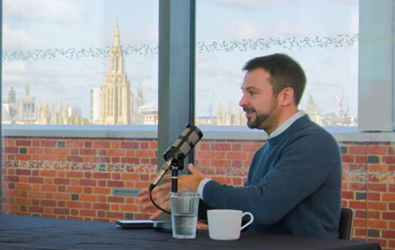 A podcast guest sits at a table talking with gesturing with their hands.