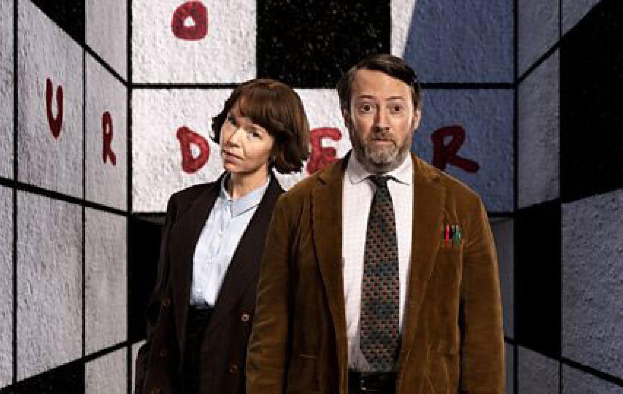Two TV characters, a man and a woman, stand in front of a crossword cover walls of a room.
