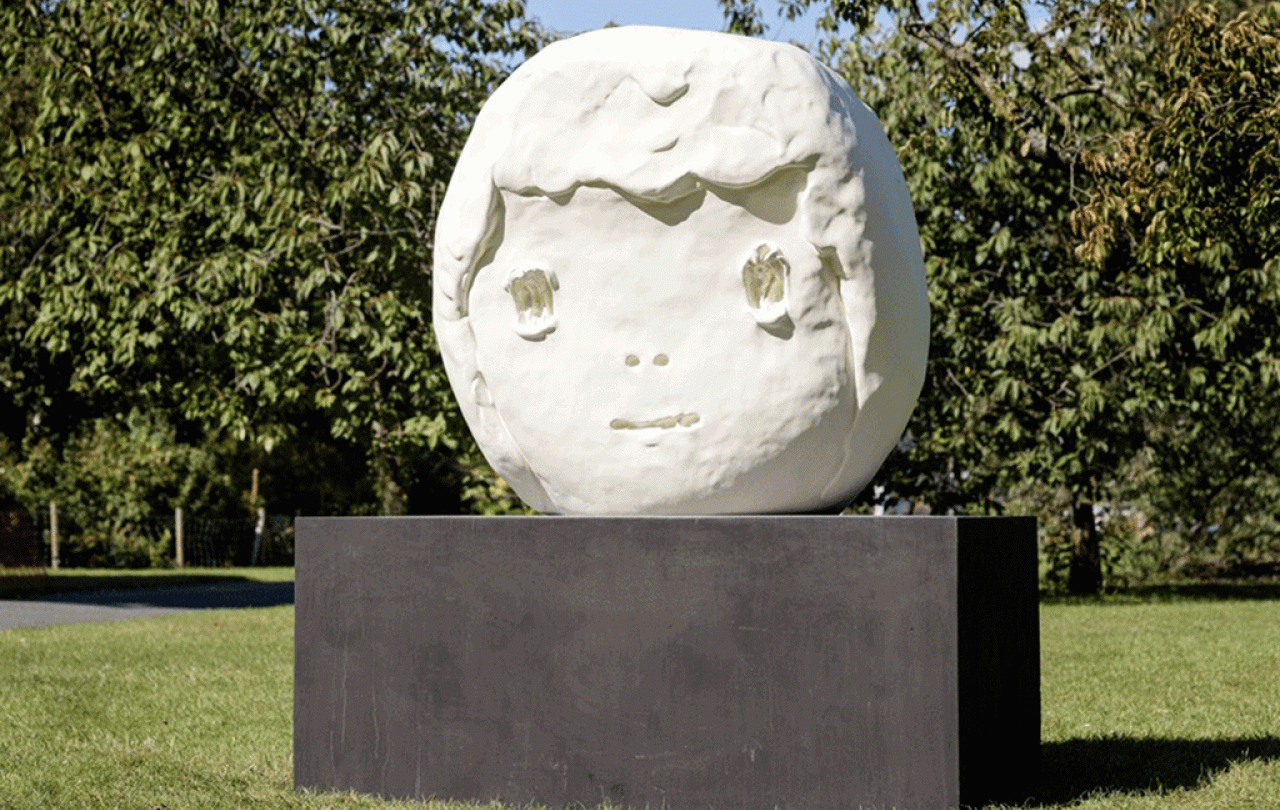 A large outdoor sculpture of a round child-like head sits on a dark plinth.