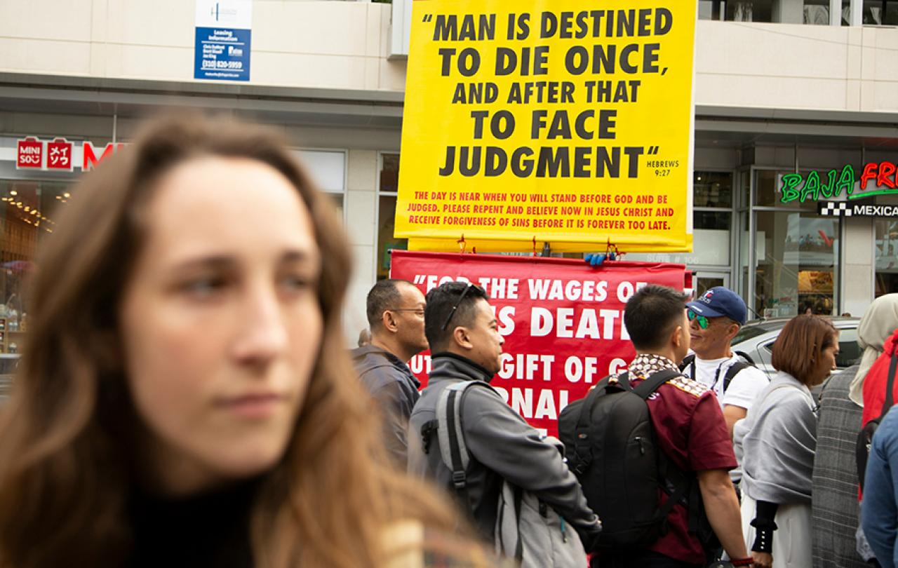 Behind a passer by a street peacher holds up a large yellow sign with a message on it.