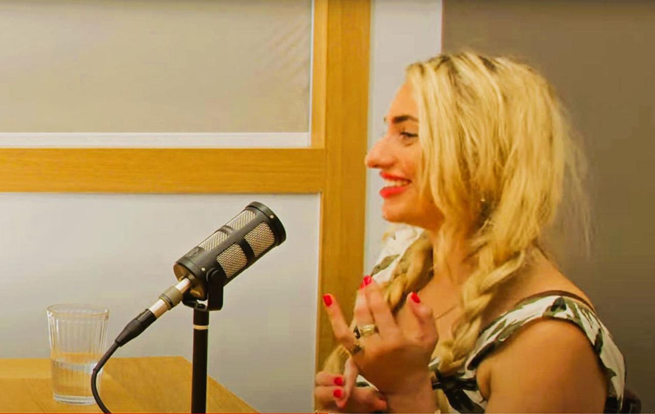  In profile a woman speaks into a microphone and holds her hands up from a desk.