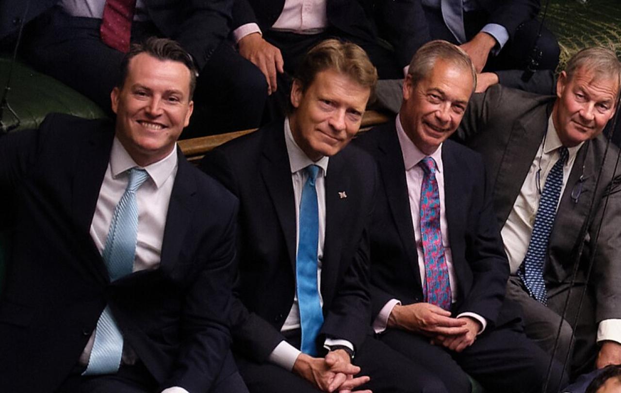 Four men in suits, sit next to each other smiling, in the House of Commons.