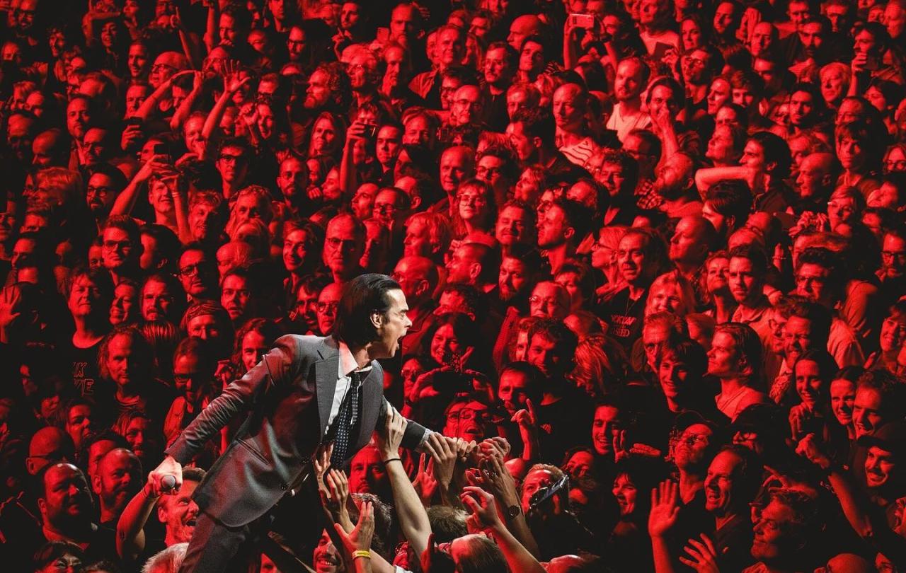 A rock star prowls the stage while behind hundred of faces tined red star.