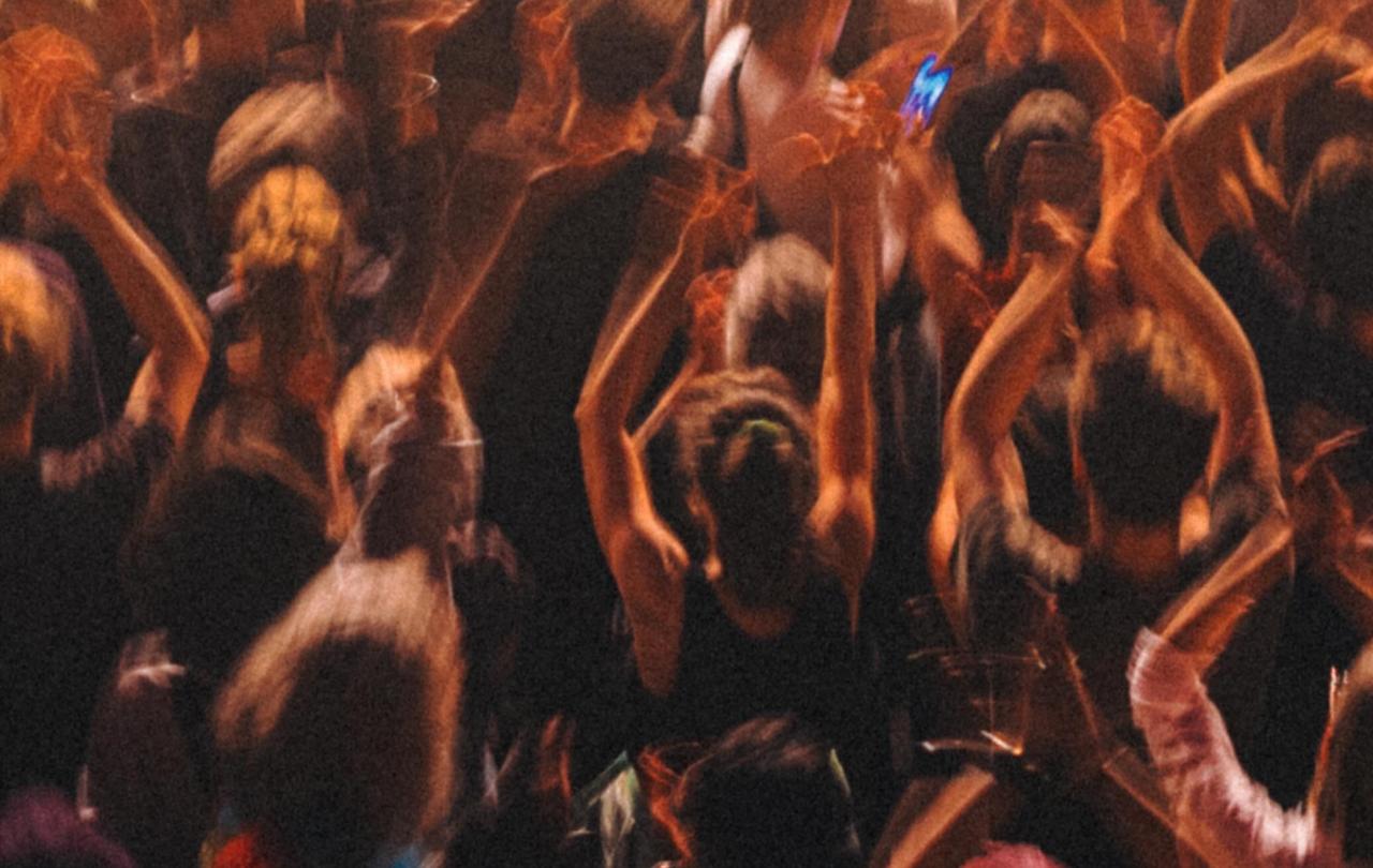 Concert goes stand, bathed in warm light, raising their hands in the air, facing away from the camera to the stage