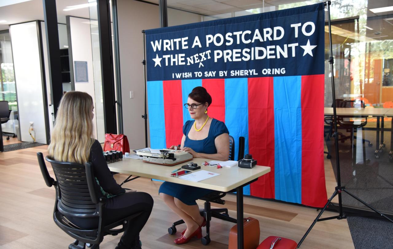 Two people sit at the table, one dictates as the other types. Behind a banner reads: write a postcard to the next president.i