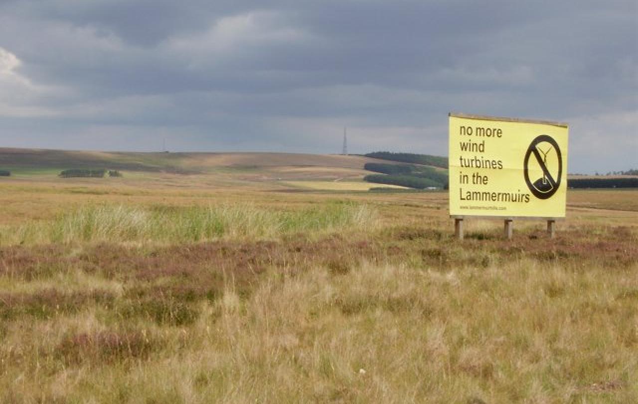 A sign protesting a proposed wind farm stands on flat moorland.