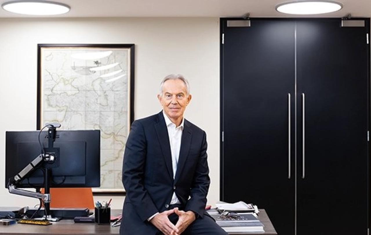 Tony Blair rests on the edge of a desk.