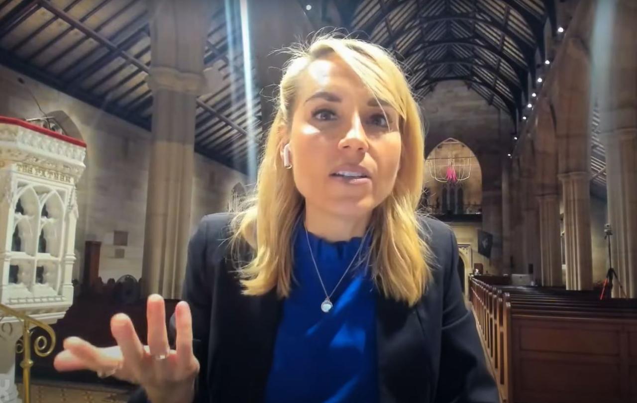 A woman sitting in an empty church talks to the camera and gestures with one hand.