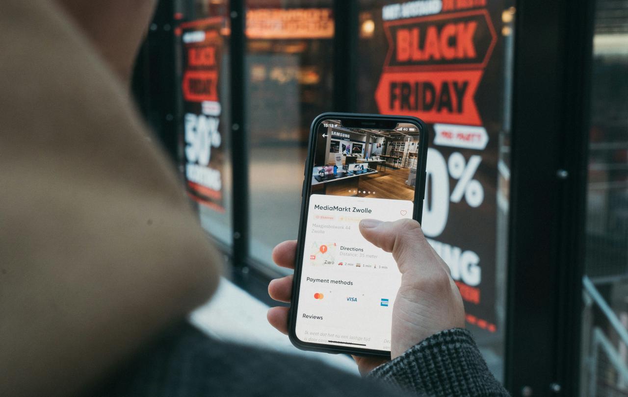 A phone screen is held up, showing a shopping page, behind a window displays a Black Friday Sale banner.