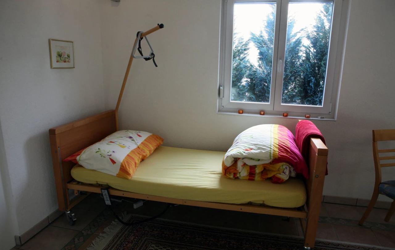 A single bed, wiith an unmade colourful duvet stands in the corner of a room. A hoist reaches over it from the corner.
