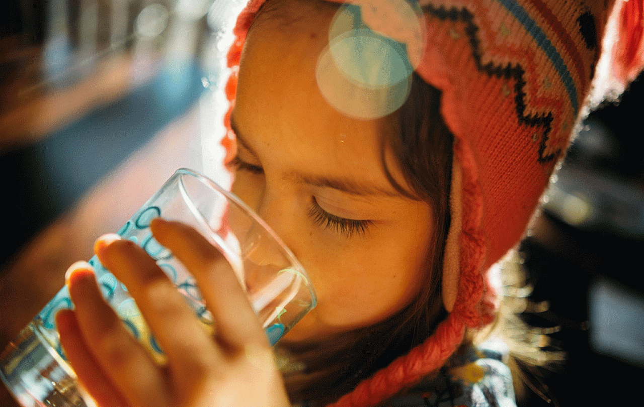 A child wearing a wool hat holds a glass and drink water from it.