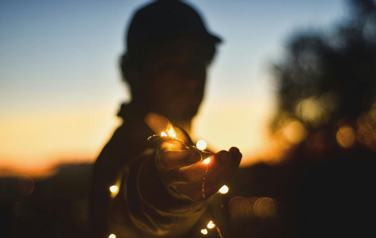 A backlit person at twilight holds a hand out towards the camera, holding some fairly lights