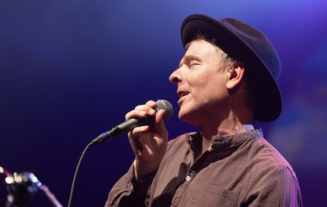 A singer, wearing a hat, pulls his head back holding a note, and a mic.