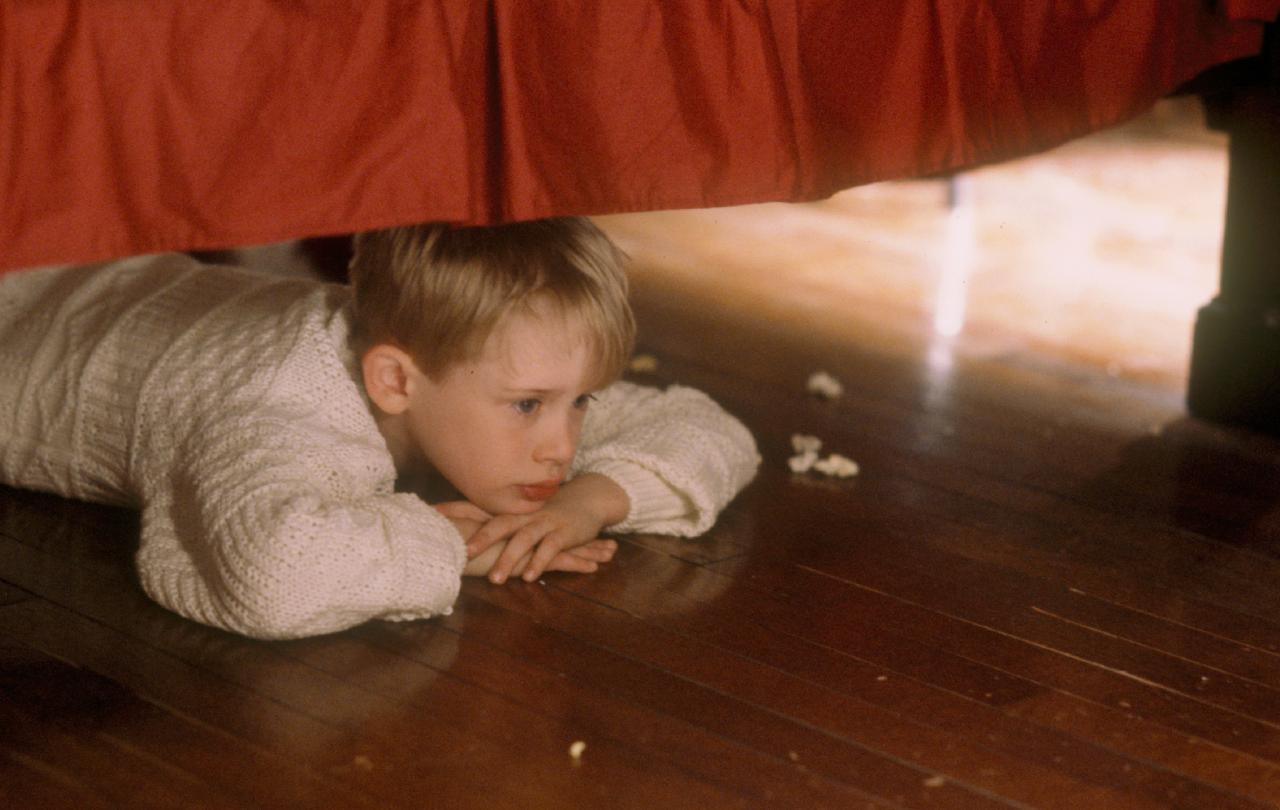 A child lies under a bed, resting his chin on his hands, contemplating.