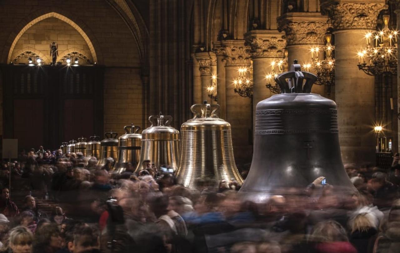 Restored church bells lined up in a cathedral, as crowds mill around them.