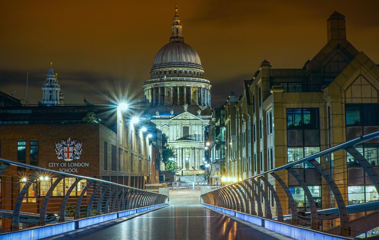 A view from a floodlight footbridge towards a gap between office buildings which reveal a cathedral and its illuminated dome.