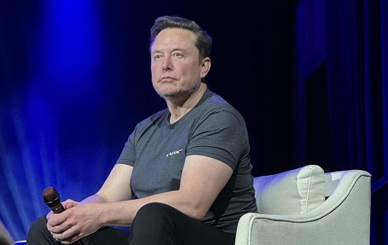 Elon Musk, wearing a t-shirt slouches forward, holding a mic, while sitting on a stage chair.